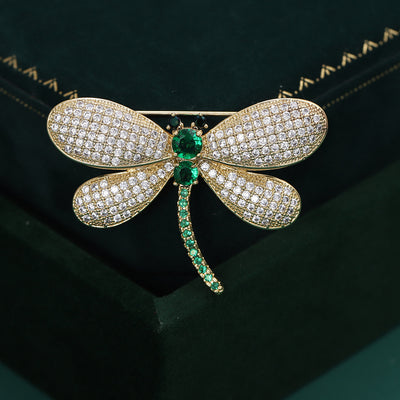 Display view of gold tone crystal dragonfly brooch with green stone body standing on white blocks.