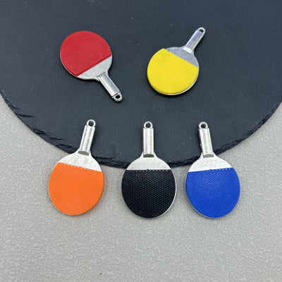 Five mini table tennis paddle charms in red, yellow, orange, black and blue arranged on a slate board