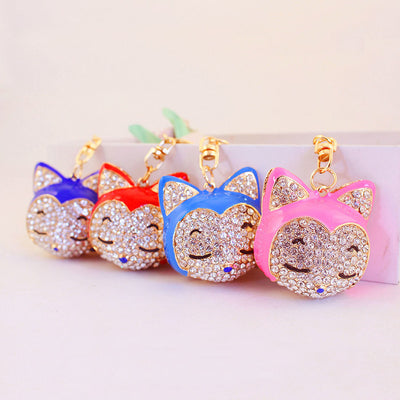 Four stone-covered cat-face keychains in pink, blue, red, and dark blue are arranged in a row.