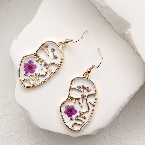 Pair of gold face-outline resin earrings with purple pressed flowers on white tray
