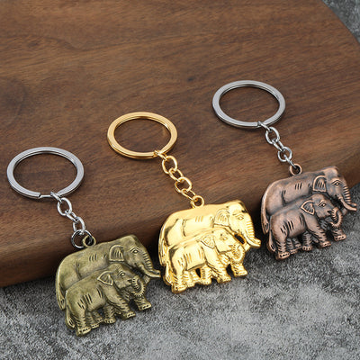 Three double-elephant keychains in gold, antique bronze, and antique copper finishes on a wood and gray surface.