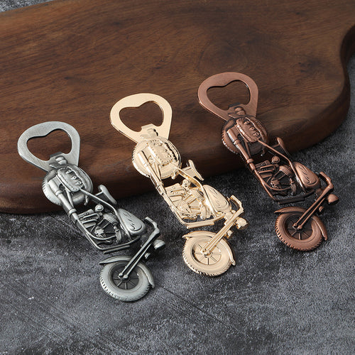 Three motorcycle-shaped openers in silver, gold, and copper finishes on dark fabric.