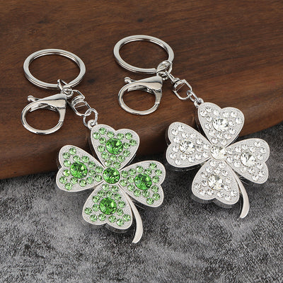 Two silver-tone clover keychains with clear and green faceted decorations rest on wood and gray fabric.