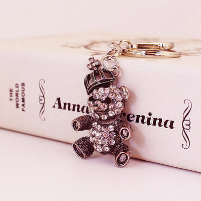 Close-up of a crowned teddy bear keychain with clear rhinestones beside a printed box.