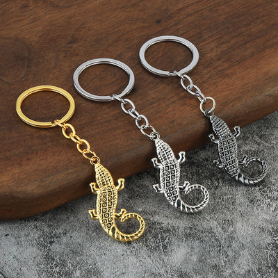 Three crocodile keychains in gold, silver, and dark metal finishes lie on a wood and gray surface.