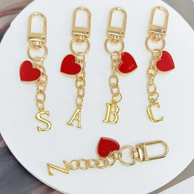 Four gold-tone letter keychains with red heart charms spelling S, A, B, C, and N on a curved white tray