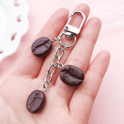 A hand holds a dark brown three-bean chain charm attached to a silver-tone snap clasp.