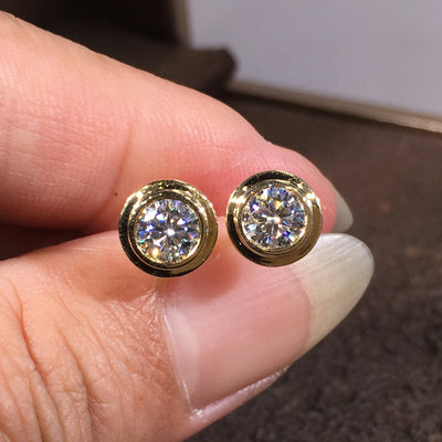 Pair of gold round bezel clear stone studs held between fingers front view