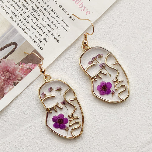 Gold face-line resin drop earrings with purple blooms beside printed book page