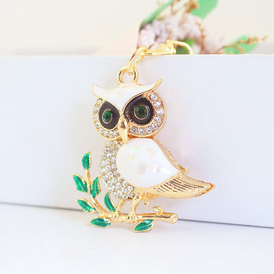 Front view of a white owl charm perched on a green leafy branch.