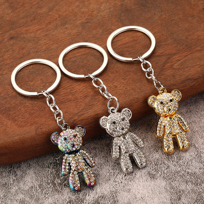 Three bear keychains in rainbow, silver, and gold tones arranged on wood and gray fabric.