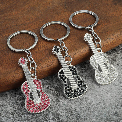Three silver-tone guitar keychains with pink, black, and clear faceted accents on a wooden surface.