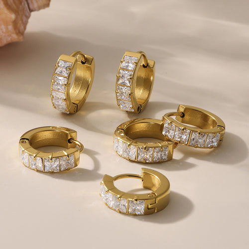Multiple gold square-zirconia huggie hoop earrings scattered on beige surface