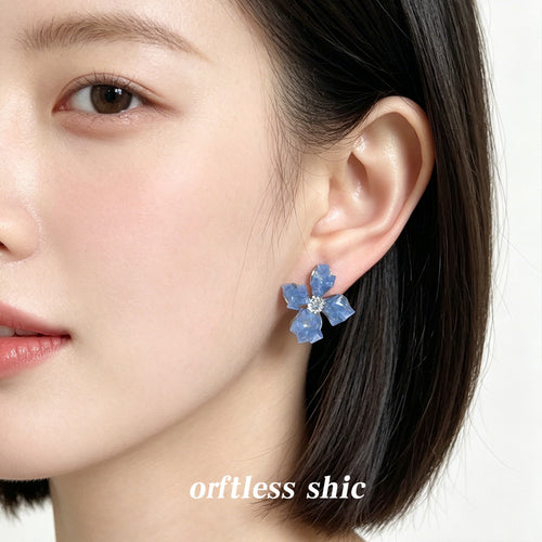 Dusty-blue translucent-four-petal flower studs shown worn on an ear.