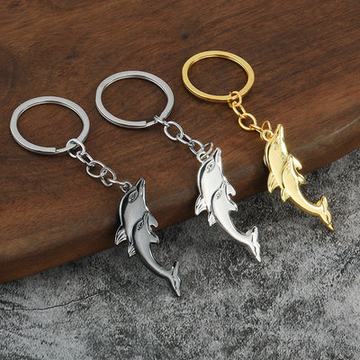 Three double-dolphin keychains in gunmetal, silver-tone, and gold-tone finishes on a wooden surface.