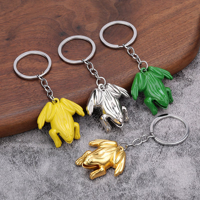 Four frog keychains in silver, gold, yellow, and green displayed on wood and gray fabric.