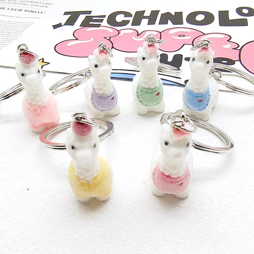 Six white flocked alpaca keychains in colored bibs and sweaters on white surface