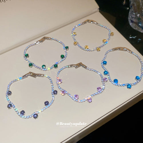 Five silver tennis bracelets with colored stone drops on cream surface