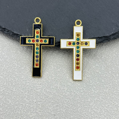 Two enamel cross charms side by side, black and white, both set with multicolor rhinestones, on a slate background