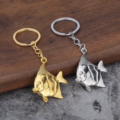 Two fish-shaped keychains in gold-tone and silver-tone finishes rest on a wood and stone surface.